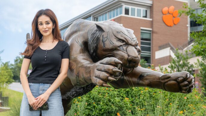 A woman with long dark hair leans on a large bronze statue of a tiger pouncing, its clawed paws reaching in front of it, with a Clemson Tiger paw on Memorial Stadium visible in the background.