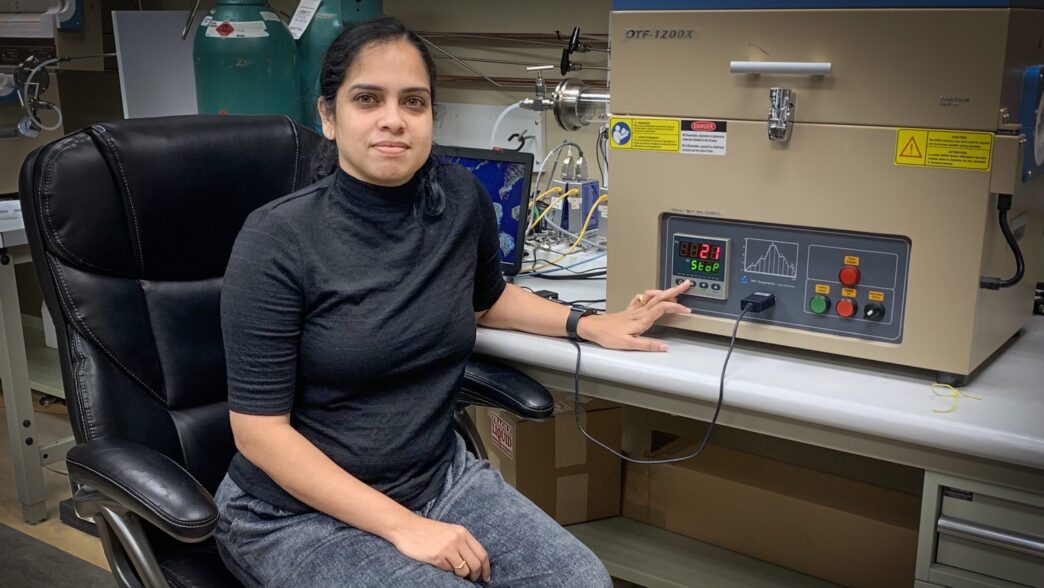 A woman wearing a black turtleneck shirt sits in an office chair in front of a desk, placing her hand on a large metal box with dials and a screen with digital numbers on its face.