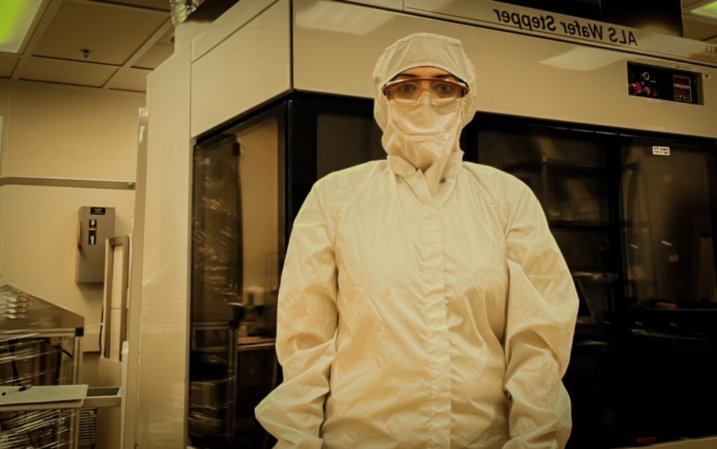 A person wearing a protective paper suit stands in a laboratory, only her eyes showing through an opening in the hood of the suit.