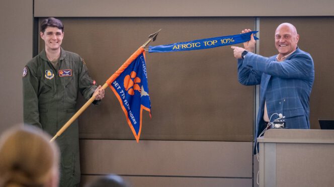 Two man stand next to each other, one holding a pole with a guidon on it and the other holding a streamer attached to the pole