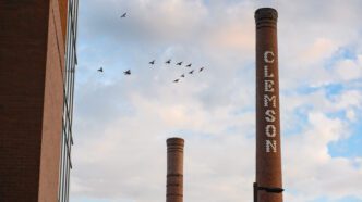 A brick smokestack with white bricks spelling Clemson down the front stands in front of a blue sky with clouds. A smaller smokestack is seen in the background.