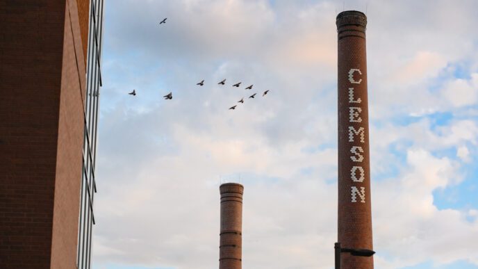 A brick smokestack with white bricks spelling Clemson down the front stands in front of a blue sky with clouds. A smaller smokestack is seen in the background.