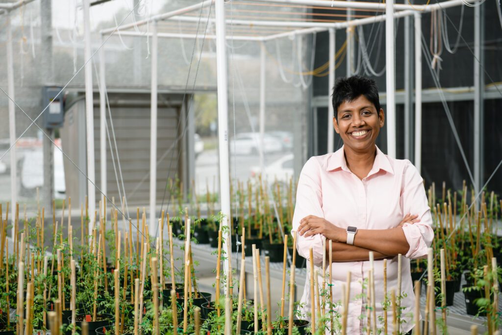 A woman with dark skin and short black hair wears a light orange shirt inside a greenhouse with small plants growing in the foreground and background.