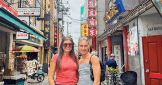 Two girls taking a picture in a street in Japan next to several stores.