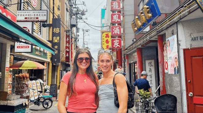 Two girls taking a picture in a street in Japan next to several stores.