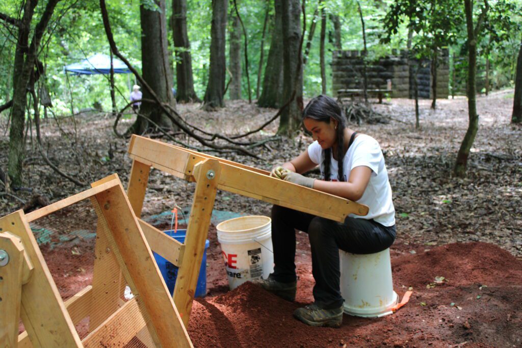 A young woman works at an easel in the woods. She has long dark braids and is sitting on top of an overturned paint bucket. 