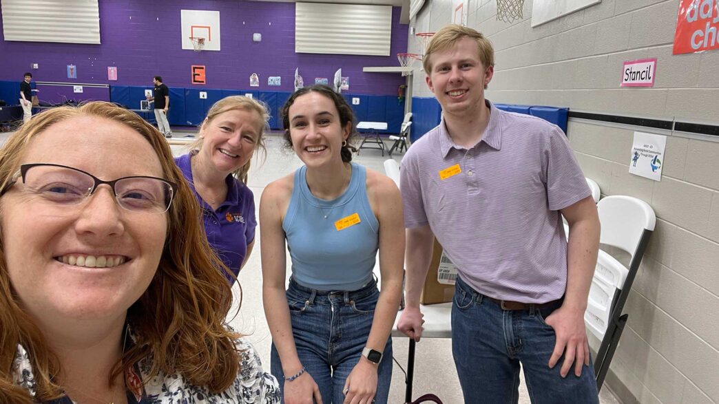 Four people standing together in a school gymnasium that is being set up for a science demonstration event.