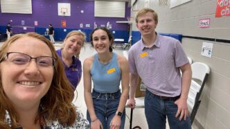 Four people standing together in a school gymnasium that is being set up for a science demonstration event.