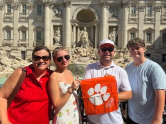 George Meister, second from right, poses with his family: wife, Emily; daughter, Taylor Leigh, and son, Jackson, in front of an old building with statues with a small Tiger Alumni rag.