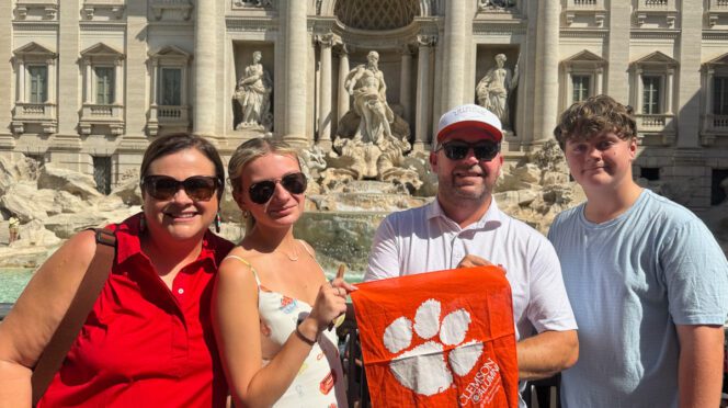 George Meister, second from right, poses with his family: wife, Emily; daughter, Taylor Leigh, and son, Jackson, in front of an old building with statues with a small Tiger Alumni rag.
