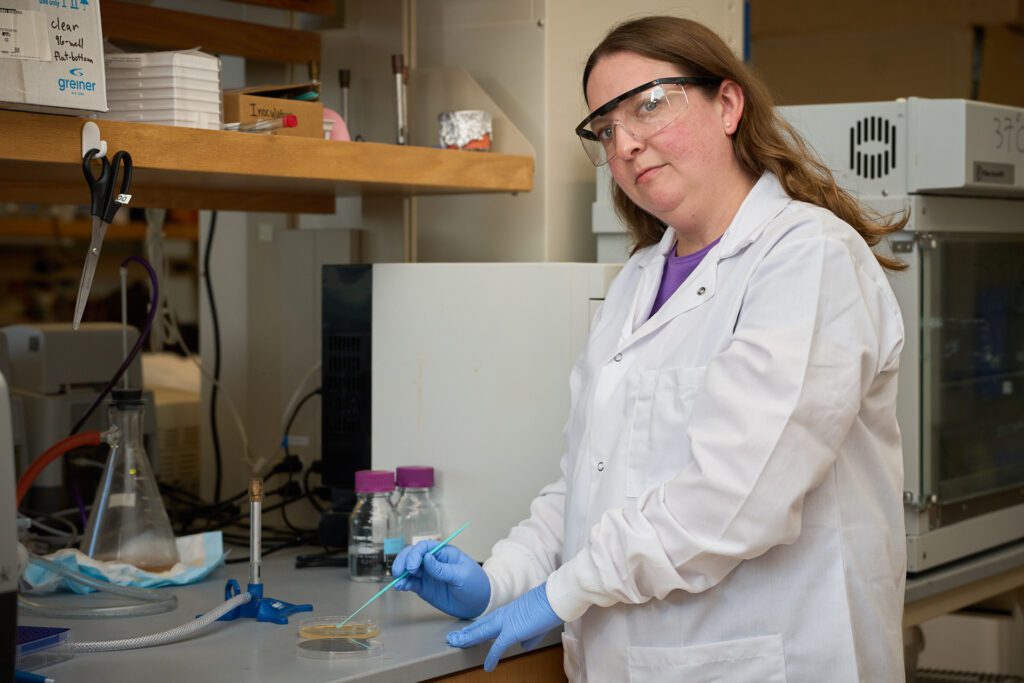 A woman wearing a white lab coat and blue gloves looks at the camera while  working with a Petri dish.