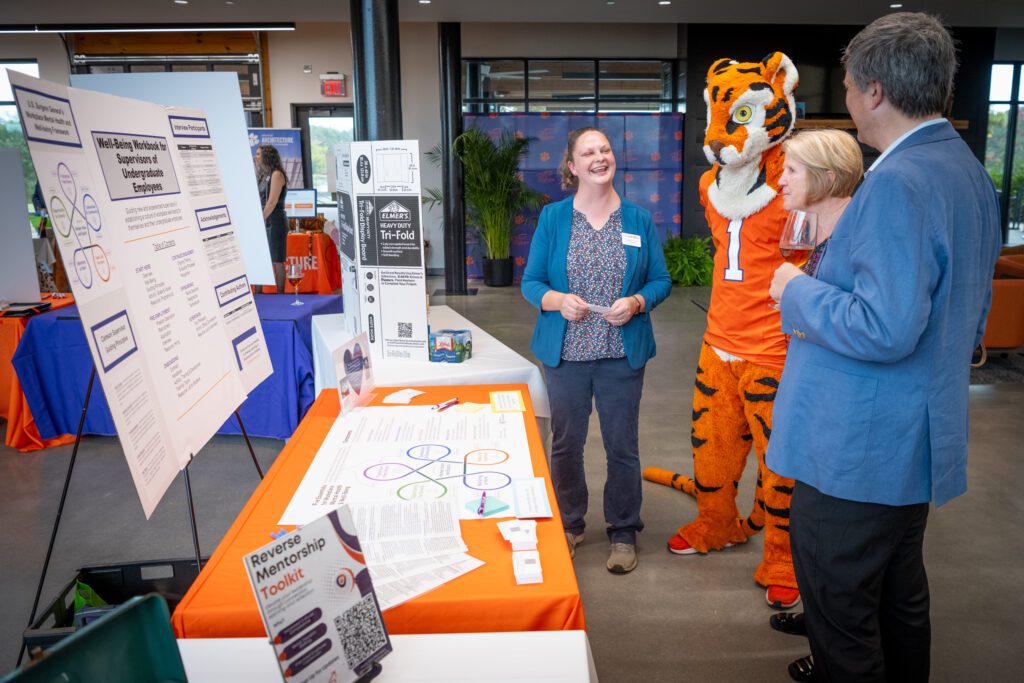 Rachel Anderson stands in front of a table with a poster presentation behind it, smiling and sharing the content with a man and a woman.