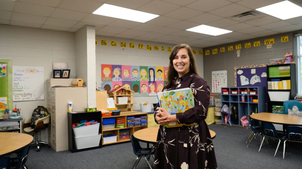 A woman with long dark hair wearing a black dress with white flowers is standing in an elementary school classroom.