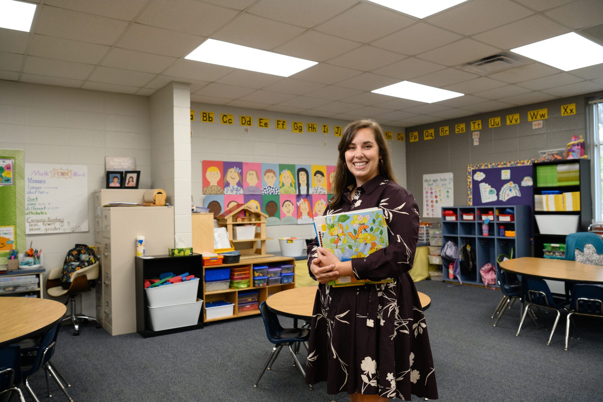 A woman with long dark hair wearing a black dress with white flowers is standing in an elementary school classroom.
