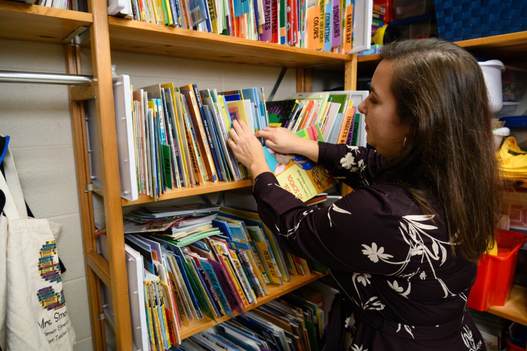 A woman with long dark hair wearing a black dress with white flowers organizes children's books on a bookshelf.