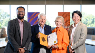 Four people stand in front of a bank of windows. Two men wearing sports coats flank and an older couple, a man with grey hair and dark blue blazer and a short-haired blonde woman wearing glasses who are holding an open portfolio that shows a resolution and Tiger Paw.