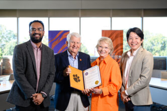 Four people stand in front of a bank of windows. Two men wearing sports coats flank and an older couple, a man with grey hair and dark blue blazer and a short-haired blonde woman wearing glasses who are holding an open portfolio that shows a resolution and Tiger Paw.
