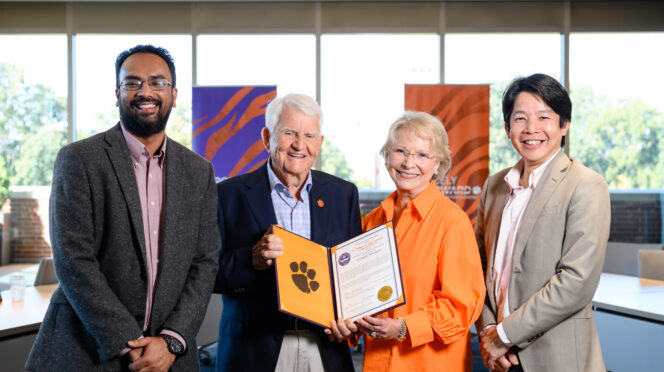 Four people stand in front of a bank of windows. Two men wearing sports coats flank and an older couple, a man with grey hair and dark blue blazer and a short-haired blonde woman wearing glasses who are holding an open portfolio that shows a resolution and Tiger Paw.