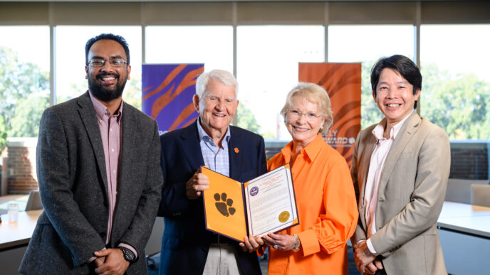 Four people stand in front of a bank of windows. Two men wearing sports coats flank and an older couple, a man with grey hair and dark blue blazer and a short-haired blonde woman wearing glasses who are holding an open portfolio that shows a resolution and Tiger Paw.