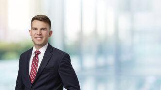 A man in a suit and red tie stands in a hall in front of out-of-focus windows.