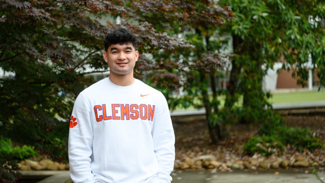 A young man with dark hair stands outside with his hands in his pockets. He is wearing a long-sleeve white shirt with Clemson emblazoned on the front.