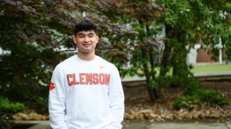 A young man with dark hair stands outside with his hands in his pockets. He is wearing a long-sleeve white shirt with Clemson emblazoned on the front.