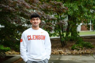 A young man with dark hair stands outside with his hands in his pockets. He is wearing a long-sleeve white shirt with Clemson emblazoned on the front.