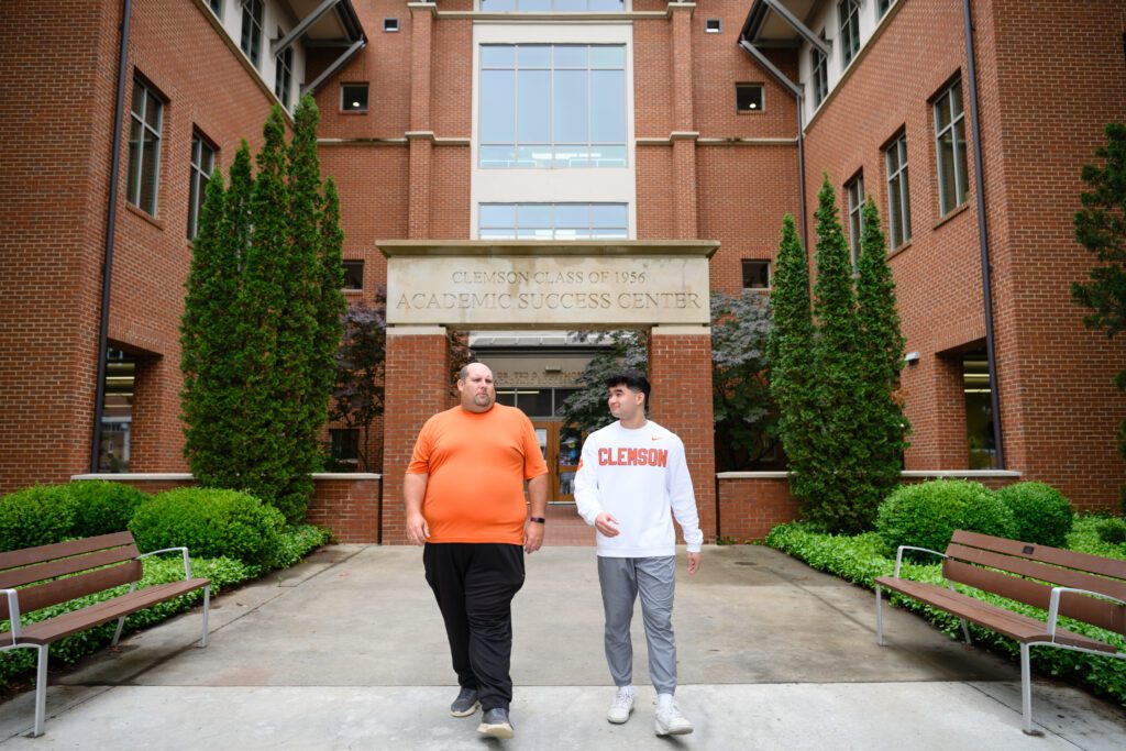 A young man on the right walks alongside an older man between two brick academic buildings.