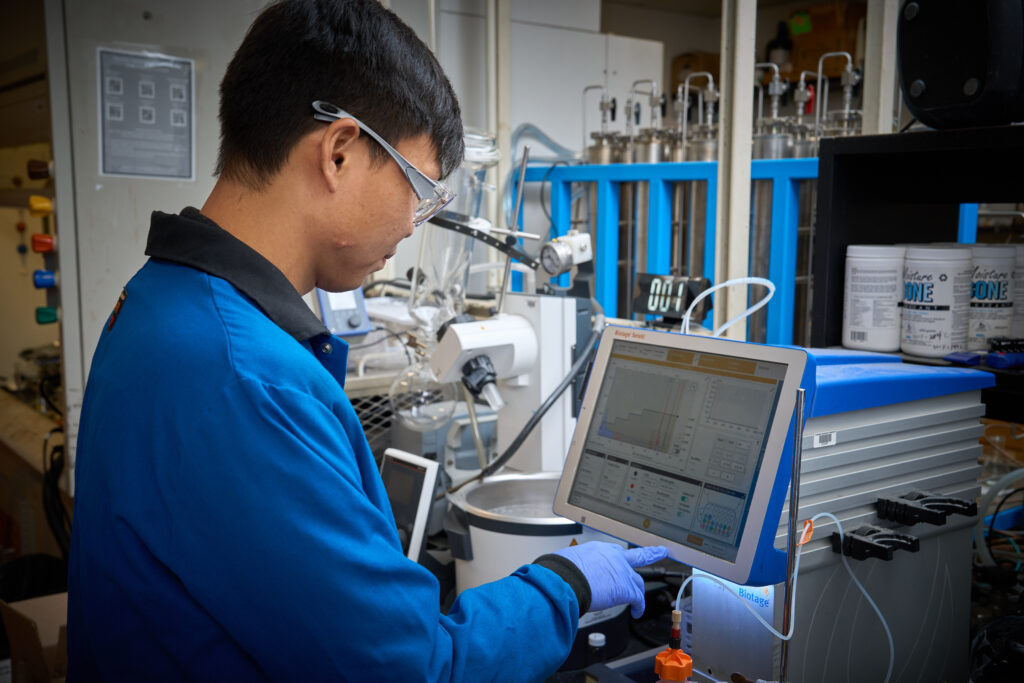 A man wearing a blue lab coat and googles is inputting information into a machine in a chemistry lab.