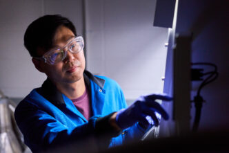 A man wearing goggles and a blue lab coat pushes a button on a machine.