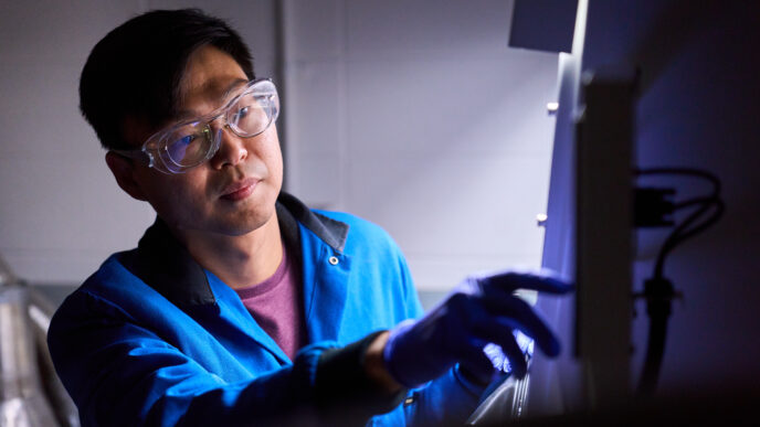 A man wearing goggles and a blue lab coat pushes a button on a machine.