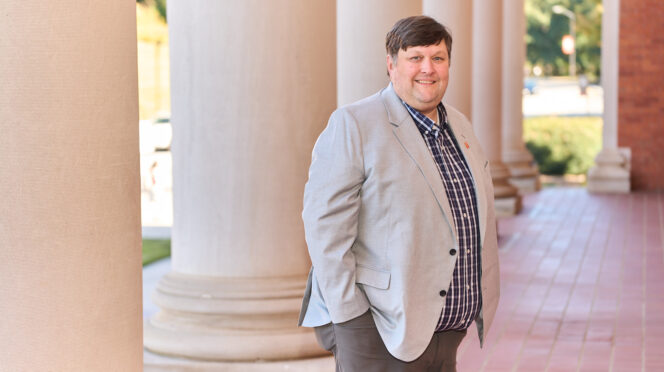 A man wearing a light colored suit stands in front of a building with columns and dark dress shirt in the background.