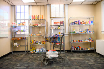 A shopping cart filled with pantry staples is positioned in front of four wire racks filled with non-perishable food items in The Nook, Clemson's employee-focused food pantry.