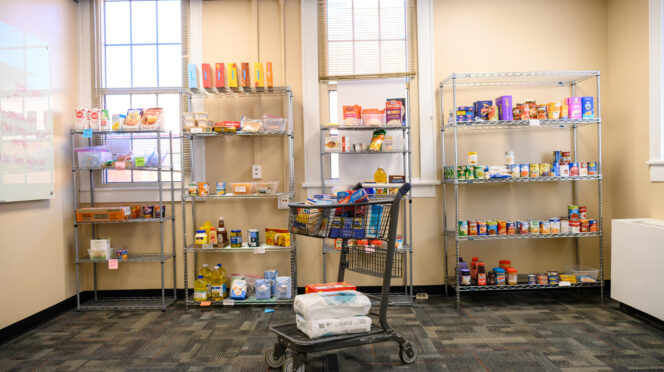 A shopping cart filled with pantry staples is positioned in front of four wire racks filled with non-perishable food items in The Nook, Clemson's employee-focused food pantry.
