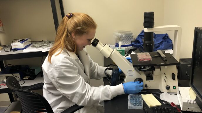 A young woman wearing a lab coat is looking through the lens of a microscope, seated in a lab.