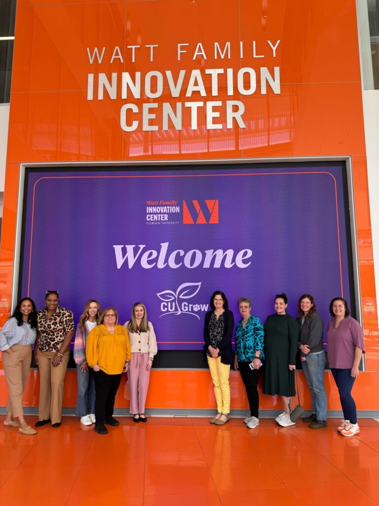 A group of Clemson staff members pose for a photo in front of a large video screen at the Watt Family Innovation Center. 