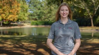 A young woman stands in front of a pond wearing a grey short-sleeve polo. She is smiling.