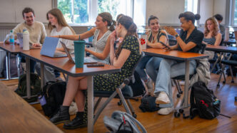Eight students sit behind desks talking and laughing with each other, with two females in the front of the room looking at a laptop.