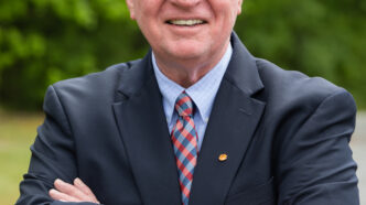 Waist up photo of an older man (Ron Gimbel) smiling, wearing a blazer and tie, arms crossed. Taken outdoors with bushes in the background.