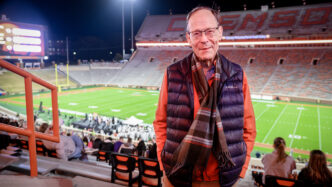 A senior gentleman dressed in a down vest and scarf stands in the bleachers of Memorial Stadium at night, the football field lit up behind him.