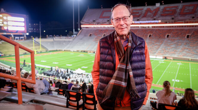 A senior gentleman dressed in a down vest and scarf stands in the bleachers of Memorial Stadium at night, the football field lit up behind him.