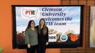 Two women stand in front of a screen that reads Clemson University welcomes PTIE team.