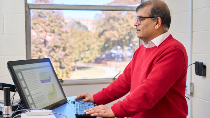 A man wearing a red sweater and white collared shirt is standing at a computer looking at the screen in a classroom.