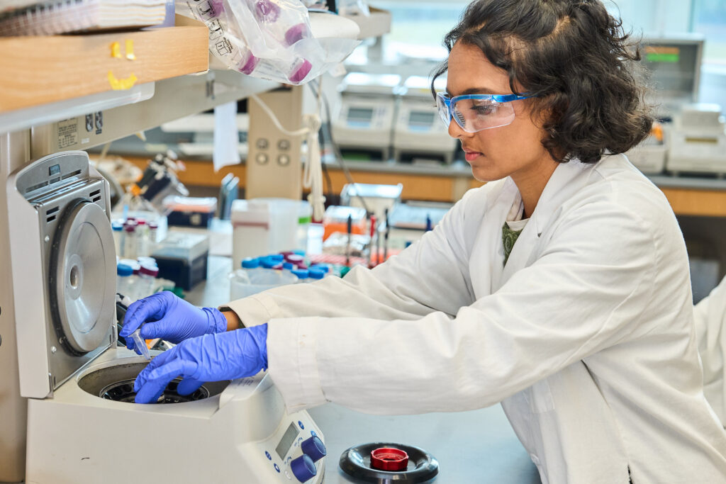 A woman with dark hair wearing safety glasses, gloves and a white lab coat puts a sample into a machine in a science lab.