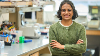 A woman with dark hair wearing a green sweater stands in a science lab with her arms crossed.
