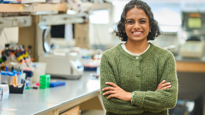 A woman with dark hair wearing a green sweater stands in a science lab with her arms crossed.