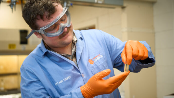 A man wearing goggles, a blue lab coat and orange gloves holds a test tube in a lab.