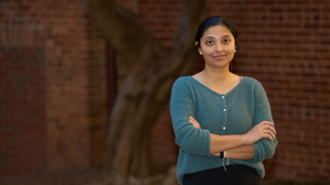 A woman wearing a green sweater stands with her arms crossed in front of a tree and building.