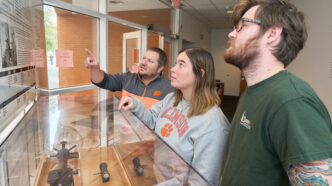 Three people look at a glass case of historic mass spectrometry equipment. Two are pointing at the display.