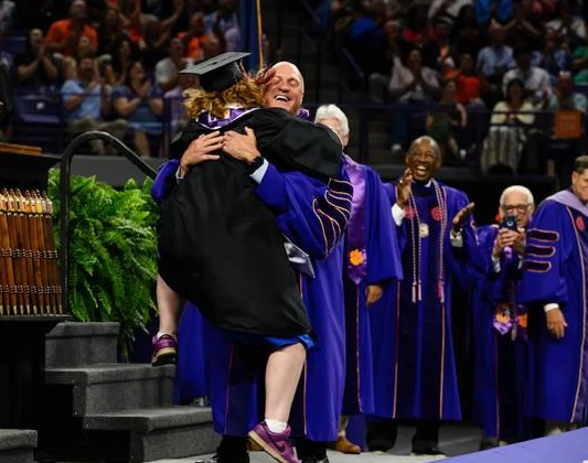 A young woman jumps into the arms of a man on a graduate stage while people look on and clap. They are all wearing academic regalia.